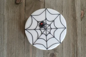 A round cookie with white icing features a black spider web design and a small black spider with a red heart on its back, ideal for a Spider Web Cookie Stamp Cookies Easy Recipe.