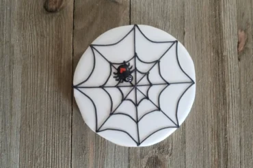 A round cookie with white icing features a black spider web design and a small black spider with a red heart on its back, ideal for a Spider Web Cookie Stamp Cookies Easy Recipe.