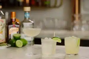 Three refreshing Lemon San Diego Cocktails served with lime garnish on a marble counter, with premium liquor bottles in the background. A perfect citrus cocktail recipe for any occasion.