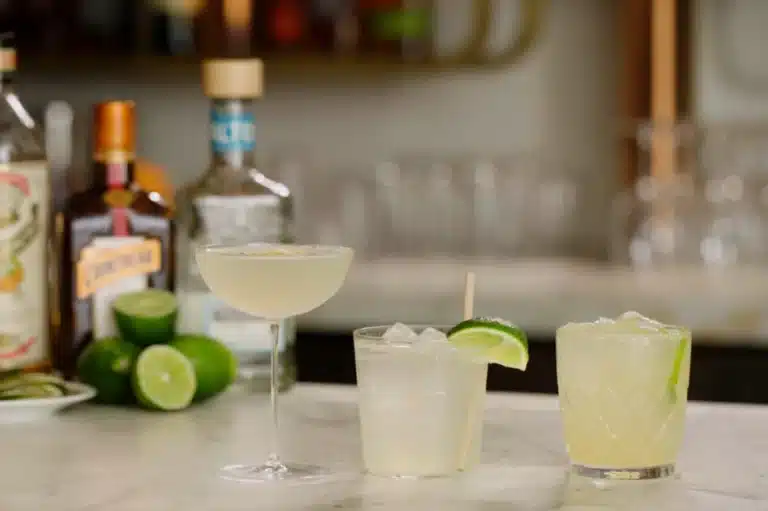 Three refreshing Lemon San Diego Cocktails served with lime garnish on a marble counter, with premium liquor bottles in the background. A perfect citrus cocktail recipe for any occasion.