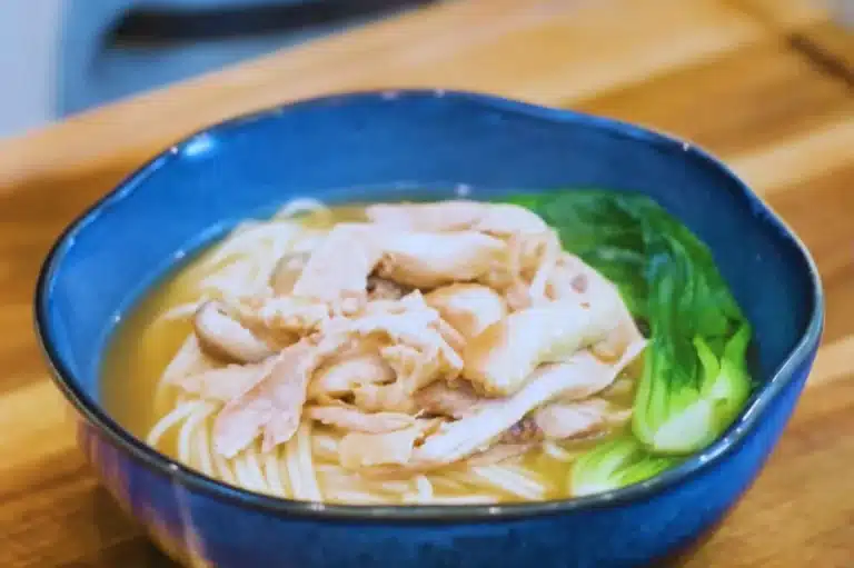 Bowl of Lemongrass Chicken Noodle Soup with shredded chicken, rice noodles, and bok choy in a rich broth served in a blue ceramic bowl.