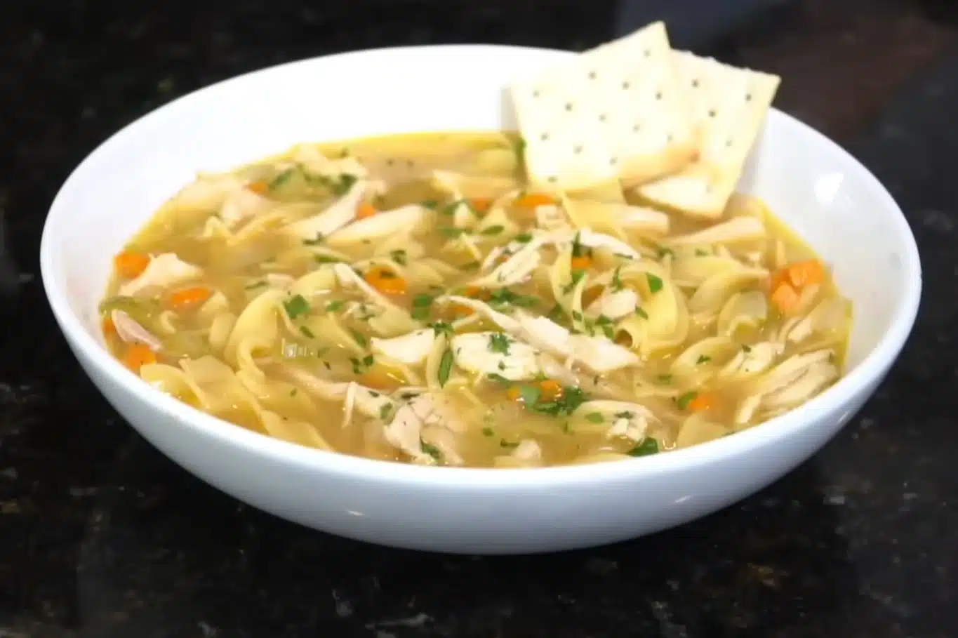 Bowl of homemade spicy chicken noodle soup with vegetables and saltine crackers on the side, served on a dark countertop