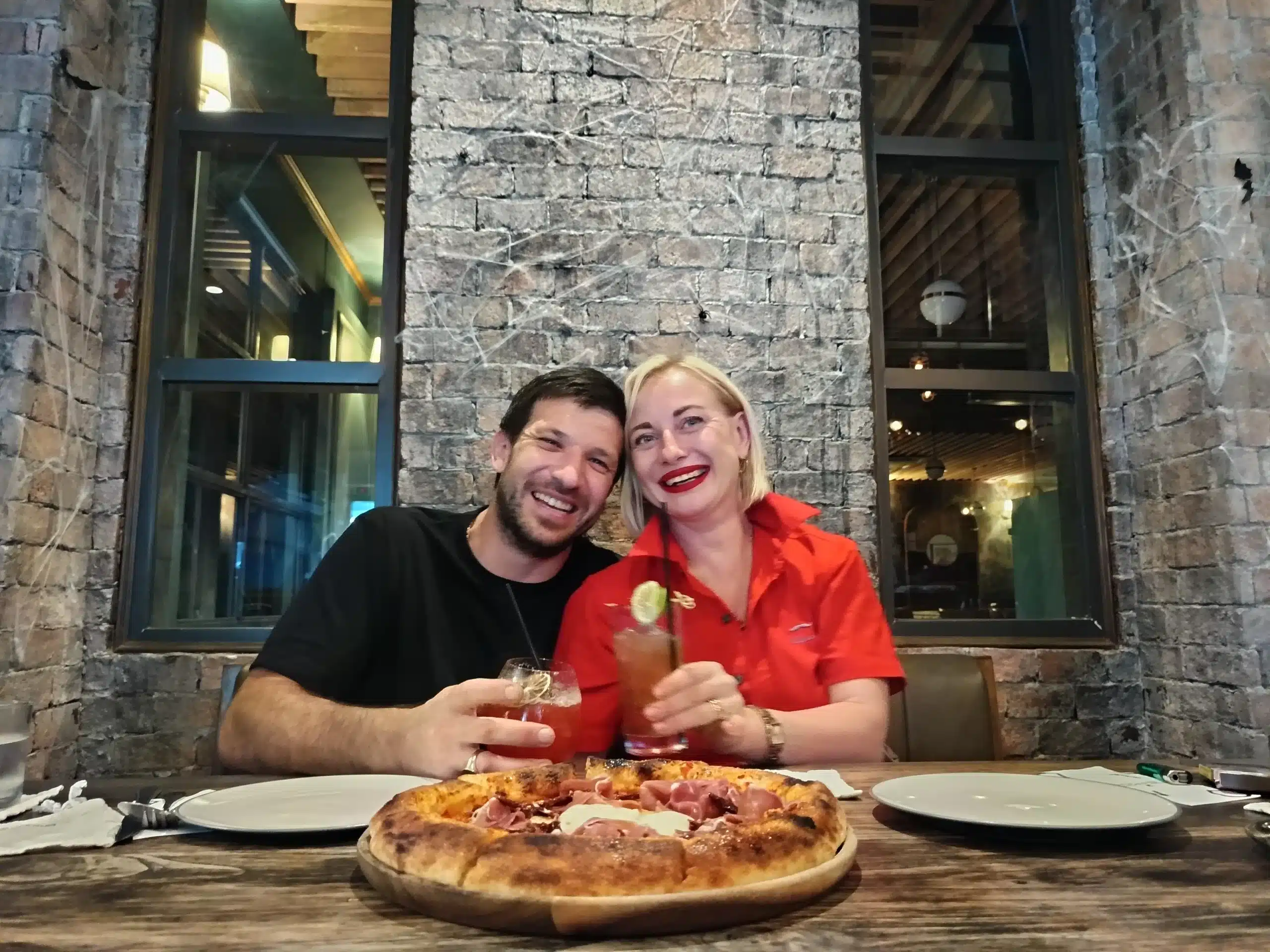 Oliver and Natalia laughing together at a table in Day & Night Restaurant with a pizza in front of them.