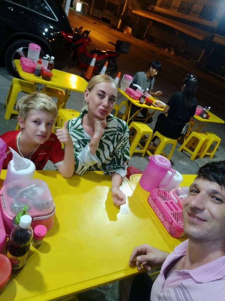 Home 1 Oliver, Natalia, and Victor at a yellow street-food table in Mae Nam, Koh Samui, before ordering ba mee kiew moo daeng