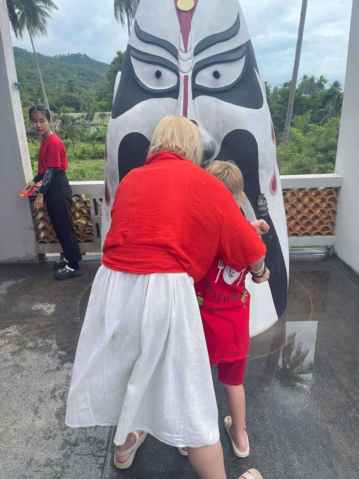 Natalia and Victor in red clothes facing a Chinese opera-mask statue on the Guan Yu shrine terrace, Hua Thanon, Koh Samui