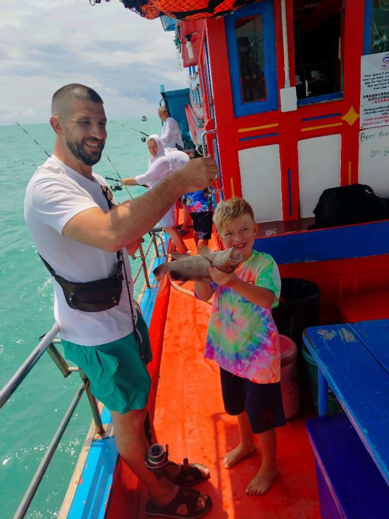 Victor holds a grouper catch beside Oliver on a red and blue Mr. Tu fishing boat, Koh Samui
