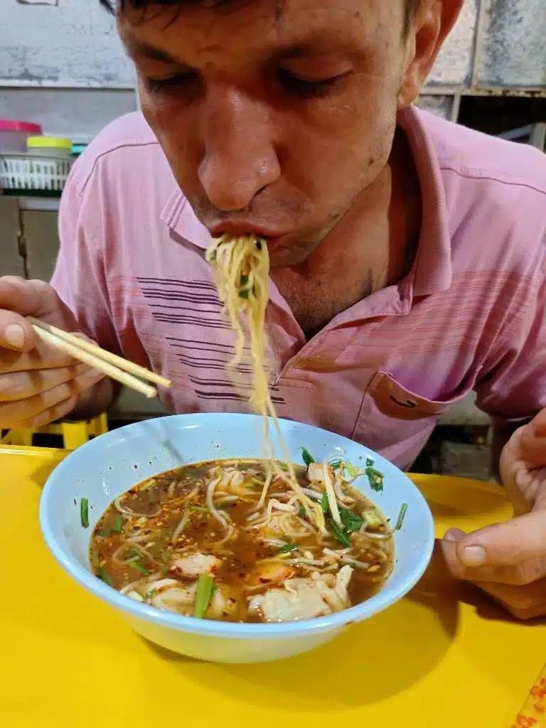 Oliver eating springy yellow ba mee noodles with chopsticks at a Thai street noodle stall